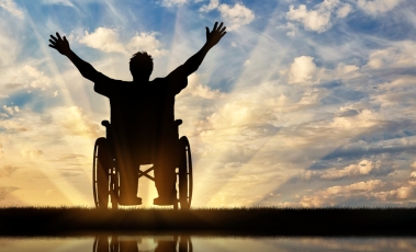 Εικόνα που δείχνει έναν άνθρωπο με αναπηρία, σε αναπηρικό αμαξίδιο, να χαιρετάει εκστατικά το ηλιοβασίλεμα.