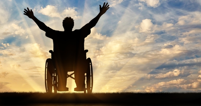 Εικόνα που δείχνει έναν άνθρωπο με αναπηρία, σε αναπηρικό αμαξίδιο, να χαιρετάει εκστατικά το ηλιοβασίλεμα.