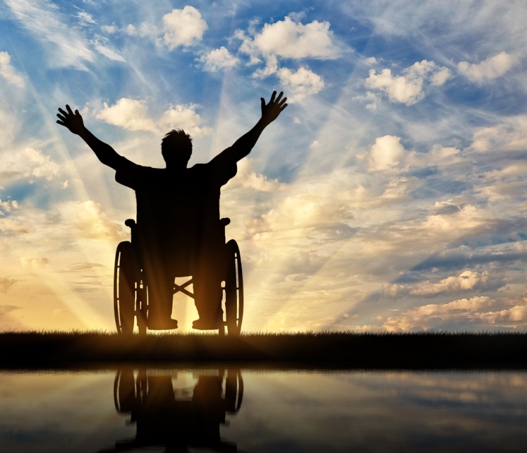 Εικόνα που δείχνει έναν άνθρωπο με αναπηρία, σε αναπηρικό αμαξίδιο, να χαιρετάει εκστατικά το ηλιοβασίλεμα.
