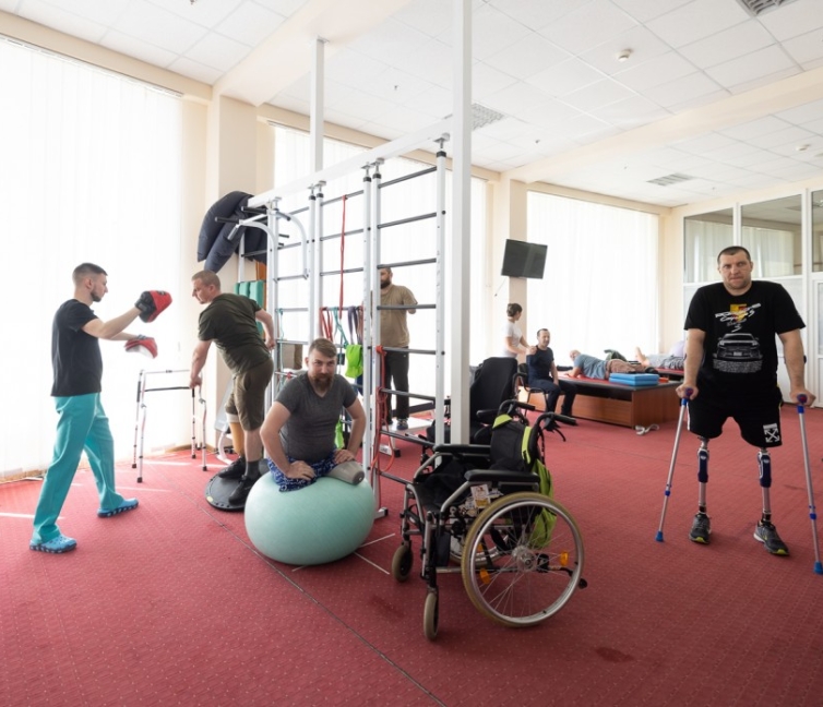 Εργαστήριο υψηλής τεχνολογίας με τεχνητά μέλη (προσθέσεις) τελευταίας γενιάς τοποθετημένα σε βάσεις. Στο βάθος διακρίνεται χώρος αποκατάστασης όπου ειδικοί βοηθούν τραυματίες πολέμου να προσαρμοστούν στη χρήση των νέων τους μελών.